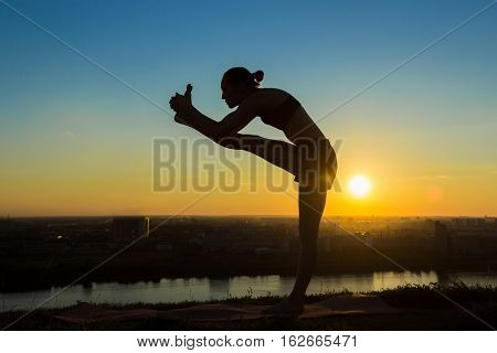 Silhouette of sporty woman practicing yoga in the park at sunset - utthita hasta padangushthasana. Sunset light, golden hour. Freedom, health and yoga concept