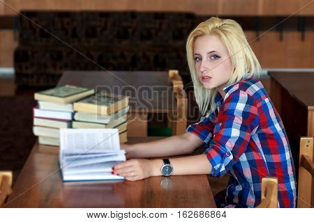 Young blonde girl in a plaid shirt sitting at desk, holding an open book in his hands. Side view.