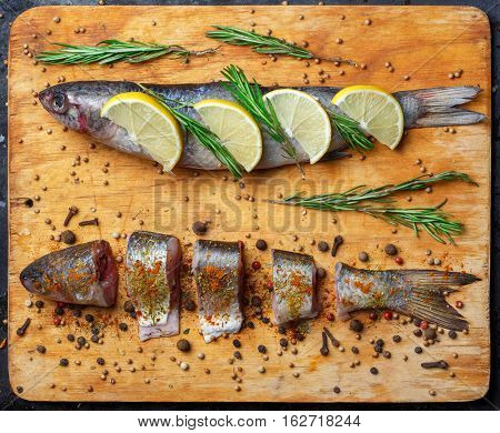 Whole and cut grey mullet fishes lie on light wooden cutting board with lemon segments rosemary branches peppercorns paprika chilli pepper and other spices around. Top view.