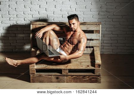 Handsome man or muscular macho bodybuilder with sexy muscle torso body with six packs and abs in sexi underpants uses laptop computer lying on wooden pallet sofa on white brick wall