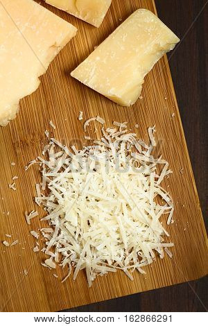 Freshly grated parmesan-like hard cheese with cheese pieces on wooden board photographed overhead with natural light