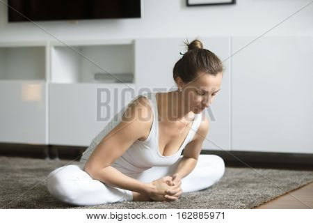 Portrait of young woman practicing yoga, sitting in Butterfly exercise, baddha konasana pose, working out, wearing white sportswear, indoor full length, home interior background