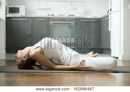 Sporty young attractive woman practicing yoga, lying in Fish exercise, Matsyasana pose, working out, wearing white sportswear, indoor full length, home interior background