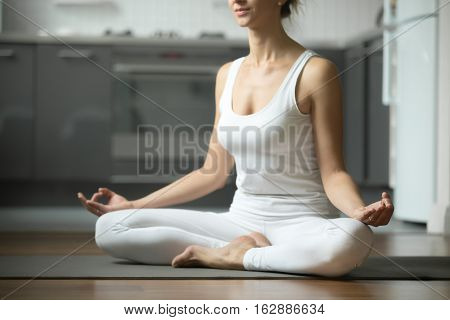 Sporty attractive woman practicing yoga, sitting in Half Lotus exercise, Siddhasana pose, working out, wearing white sportswear, indoor, home interior background. Midsection close up