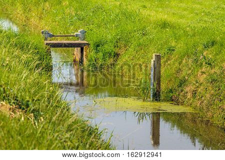 Blue tide gauge reflected in the mirror smooth water surface of a stream and in the background a simple adjustable weir.