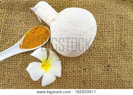 Spa herbal compressing ball white frangipani flowers (Plumeria spp Apocynaceae Pagoda tree Temple tree) and turmeric powder in wooden spoon on brown sack fabric background