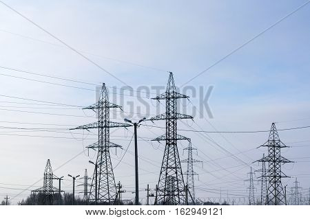 Several metal electricity pylon in the field. Cloud blue sky slight haze. The supports holding the wire. Visible tops of the trees.