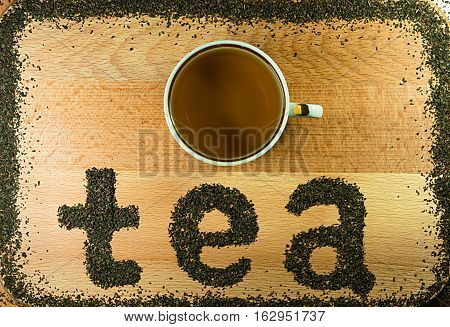 The word tea is laid out on a light-brown wooden Board small leaf black tea. Board is the background. The edges lined with a frame of tea. On Board is a mug.
