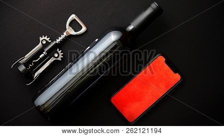 Red wine bottle, corkscrew and modern smartphone with red wallpeper on screen on black matte background. Top view with copy space