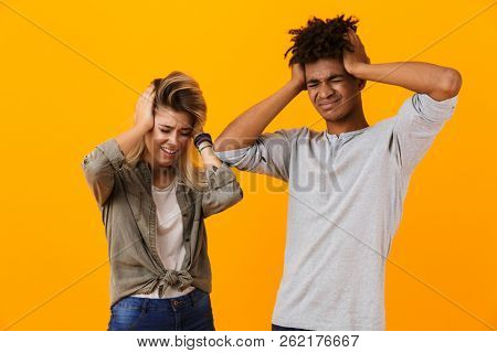 Portrait of a multiethnic couple standing isolated over yellow background, covering ears with hands