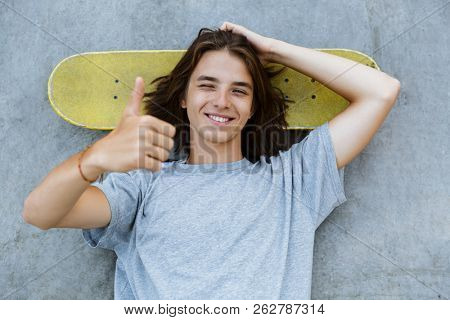 Top view of a smiling young teenge boy spending time at the skate park, laying on a skateboard at a ramp, showing thumbs up