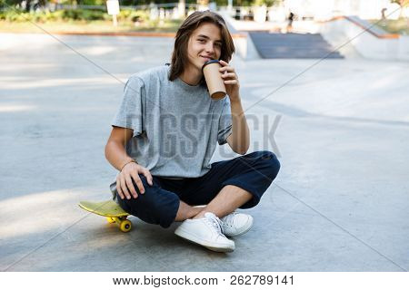 Happy young teenge boy spending time at the skate park, sitting on a skateboard, holding takeaway coffee