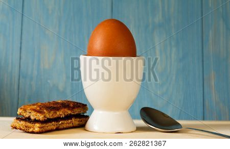Egg And Soldiers. An Egg In A White Egg Cup With Toast Soldiers With A Spoon Against A Blue Wooden B