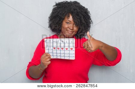 Young african american woman over grey grunge wall holding menstruation calendar happy with big smile doing ok sign, thumb up with fingers, excellent sign