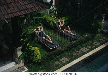 Two Young Women Lying On Lounge Chairs On Terrace Of A Tropical Hotel