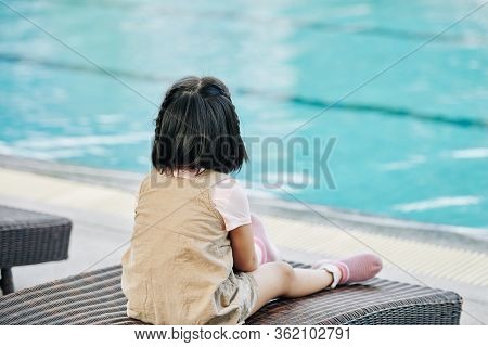 Pensive Little Girl Sitting On Chaise-lounge And Looking At Turquoise Water In Swimming Pool
