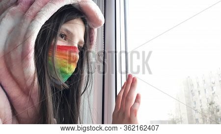 Brunette Teenager Girl In A Medical Mask Painted In Bright Rainbow Colors Stands At Window With Her 