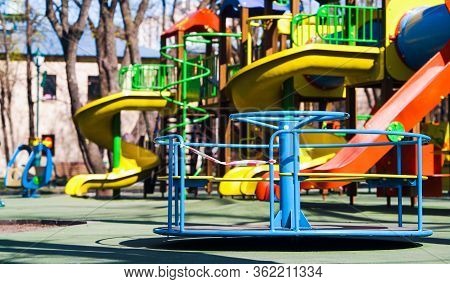 Carousel With Restriction Tape Spinning On Empty Playground In Public Park, Colorful Slides On Backg
