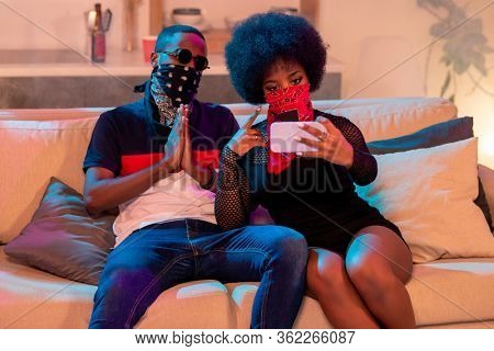 Young African man in black face bandana keeping hands together by chest while sitting next to girl with red nikab making selfie