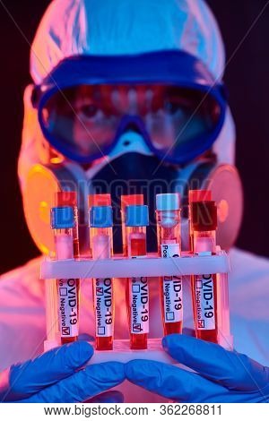 COVID-19. Close-up portrait of a doctor in a protective suit showing blood samples of infected people in test-tubes. Coronavirus test. Black background with red and blue light.