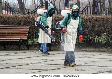Workers In Chemical Protection Suits Decontaminate A City Street Or Park During Quarantine And A Pan