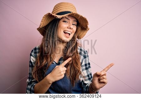 Young beautiful brunette farmer woman wearing apron and hat over pink background smiling and looking at the camera pointing with two hands and fingers to the side.