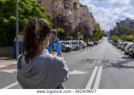 A Young Girl In Protective Gloves And A Mask Photographs An Empty Street On A Mobile Phone. No Peopl
