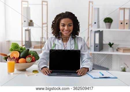 Happy African Woman Dietologist Showing Blank Laptop Screen, Sitting At Her Cabinet, Nutritionist Co