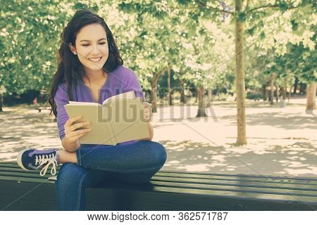 Beautiful Smiling Student Reading Book In Park. Attractive Young Woman Sitting On Wooden Bench In Pa