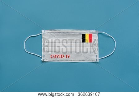Medical Mask With The Flag Of Belgium And The Inscription Covid-19 On A Blue Background
