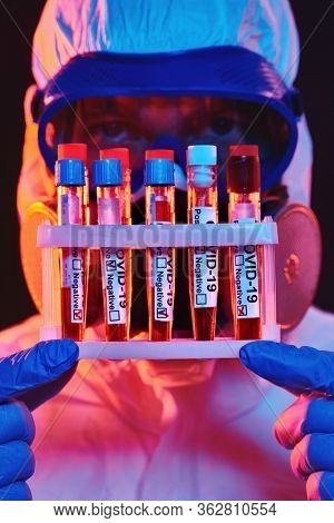 COVID-19. Close-up portrait of a doctor in a protective suit showing blood samples of infected people in test-tubes. Coronavirus test. Black background with red and blue light.