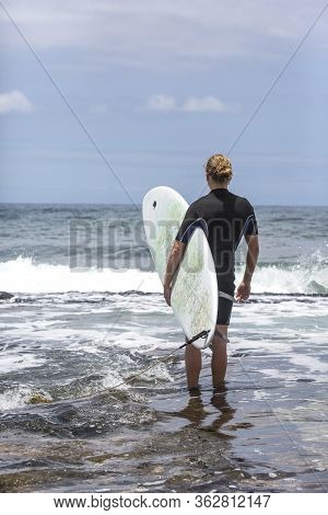 The Surf Enters The Water. Male Surfer Entering The Sea With His Board In A Black Surfing Suit. Tene
