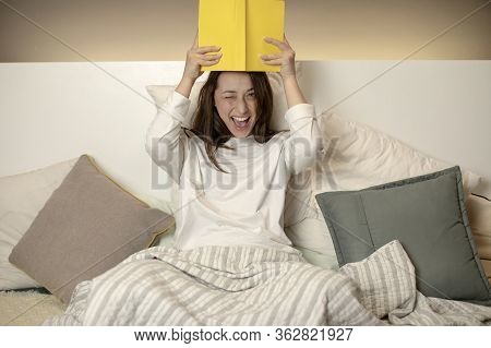 Joyful Beautiful Smiling Girl In Casual Wear Sitting In Bed, Holding Book Over Head And Playfully Wi