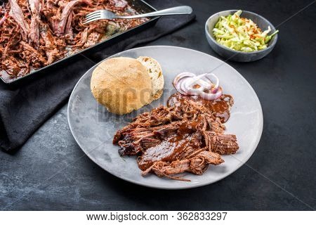 Traditional barbecue wagyu pulled beef offered with Carolina BBQ sauce, onion rings and a bun as closeup on a modern design plate 