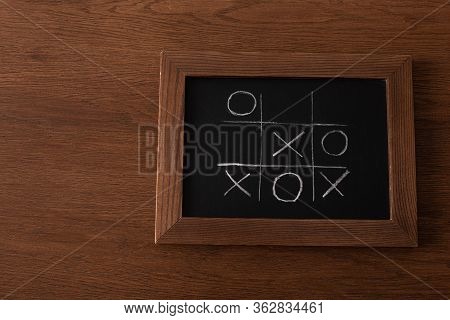 Top View Of Tic Tac Toe Game On Blackboard With Chalk Grid, Naughts And Crosses On Wooden Surface