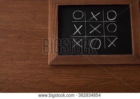 Top View Of Tic Tac Toe Game On Blackboard With Chalk Grid, Naughts And Crosses On Wooden Surface