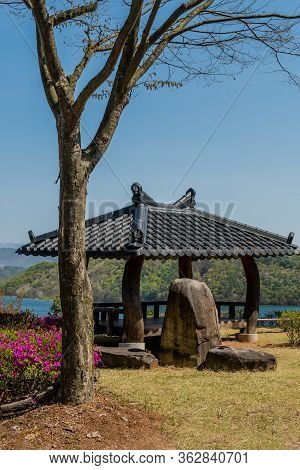 Jinan, South Korea; April 21, 2020: Pavilion With Ceramic Tiled Roof Behind Boulders And Leafless Tr