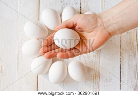 A White Chicken Egg Lying In A Man’s Hand, Shot On A White Painted Wooden Surface. Background For Li