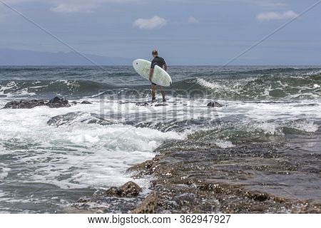 The Surf Enters The Water. Male Surfer Entering The Sea With His Board In A Black Surfing Suit. Tene