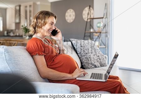 Pregnant mid adult woman using laptop and talking with a client on mobile phone. Happy expectant businesswoman sitting on couch at home in smart working. Smiling woman in maternity working on computer