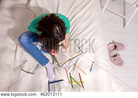 A Girl In A Green T-shirt And Blue Jeans Is Sitting On The Bed And Writing In A Notebook, Colored Pe