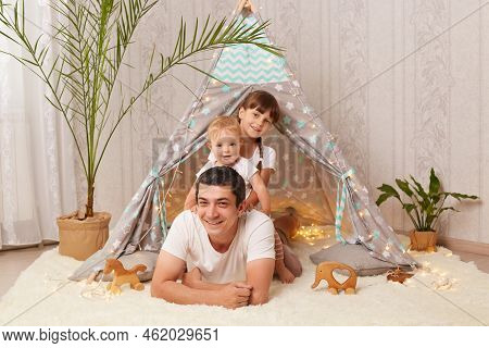Indoor Shot Of Brunette Satisfied Caucasian Man Wearing White T Shirt Lying On Floor With His Little