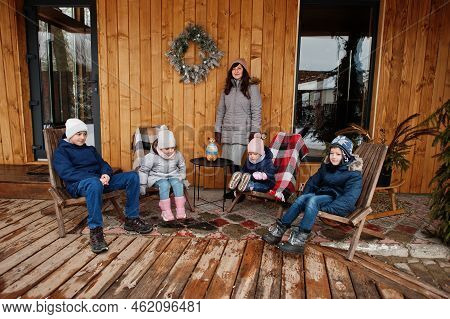 Attractive Mother With Her Four Kids On The Terrace Of A Wooden House  In Winter Day.