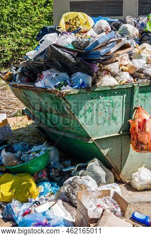 Overfilled Trash Dumpster In Ghetto Neigborhood. Trash Container Surrounded By Heaps Of Garbage.