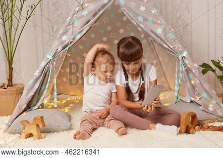 Indoor Shot Of Adorable Little Girls Wearing White T Shirts Sitting On Floor In Wigwam And Looking A
