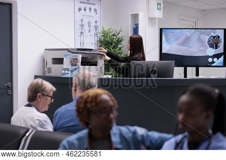 Asian Receptionist Working With Copy Printer At Hopital Reception Desk To Do Medical Reports And Reg