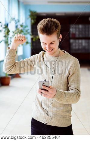 Happy Male Student Rises His Hand Up While Looking To Mobile Phone. Cheerful Student Received Messag