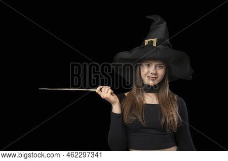 Porttrait Of Young Witch In Pointed Hat. Magic Wand In Hands. Copy Space For Text. Isolated On Black