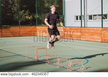 Blond Boy In Sportswear Jumps Over Red Obstacles To Improve Lower Body Dynamics. Plyometric Training