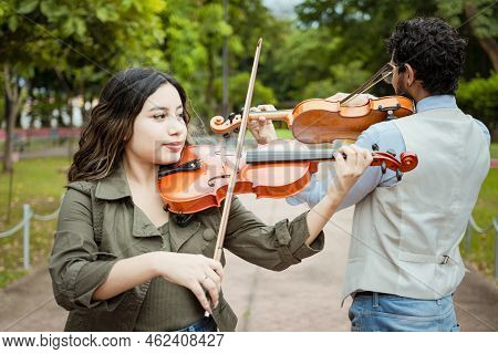 Two Young Violinists Standing Playing Violin In A Park. Portrait Of Man And Woman Together Playing V